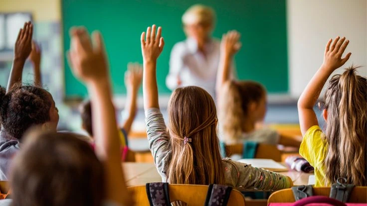 Maestra haciendo una pregunta y alumnos con manos levantadas